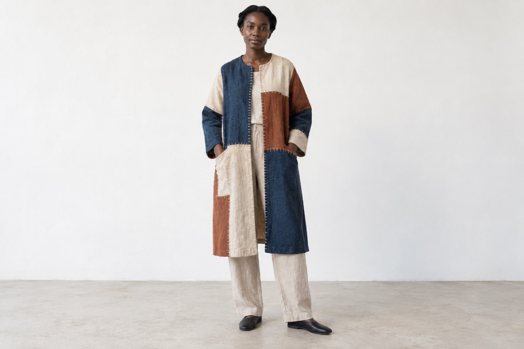 A model wears a linen, indigo and rust patchwork coat with visible hand stitching.