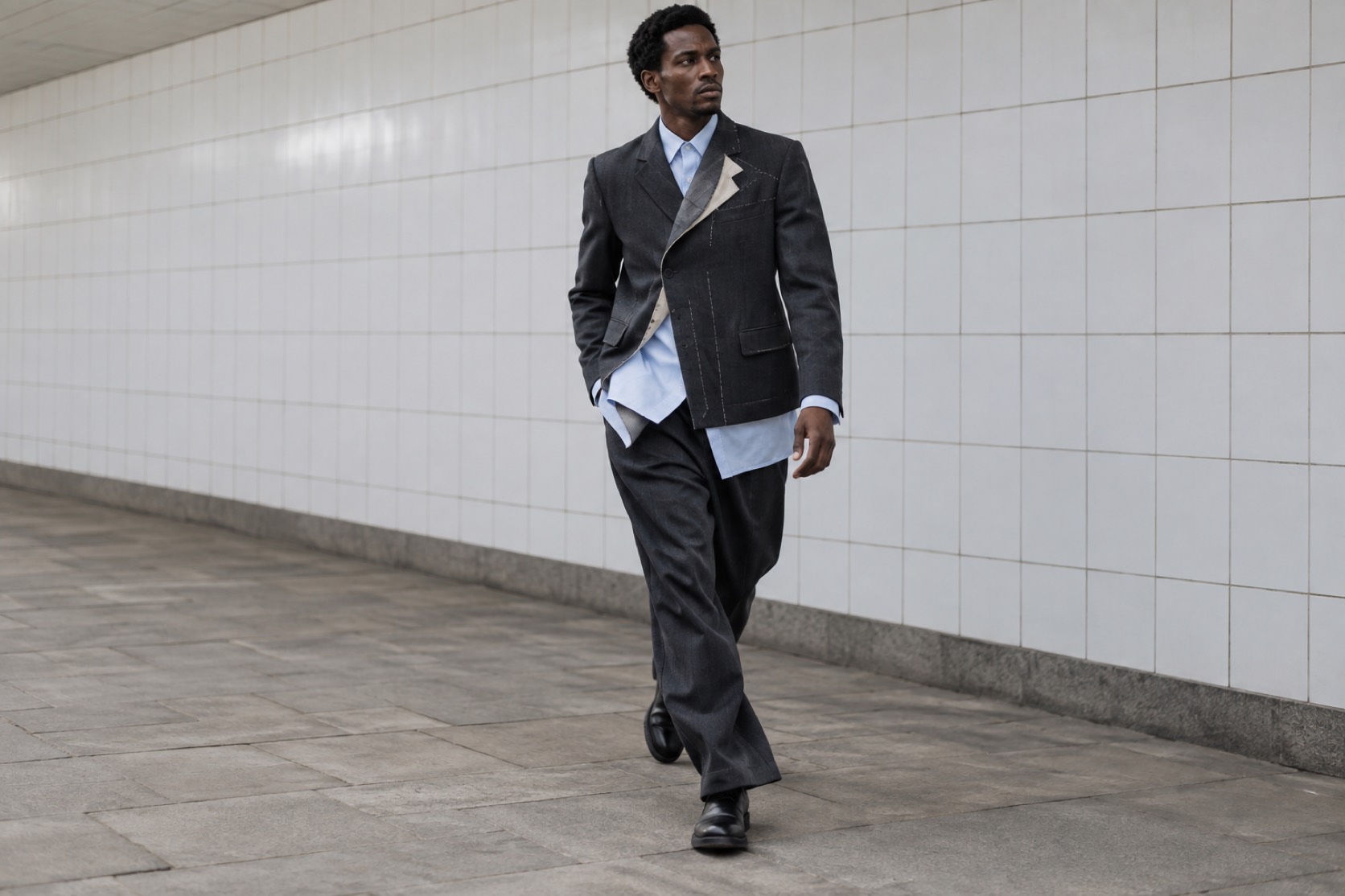 A model walks through a tiled passage in asymmetric charcoal tailoring.