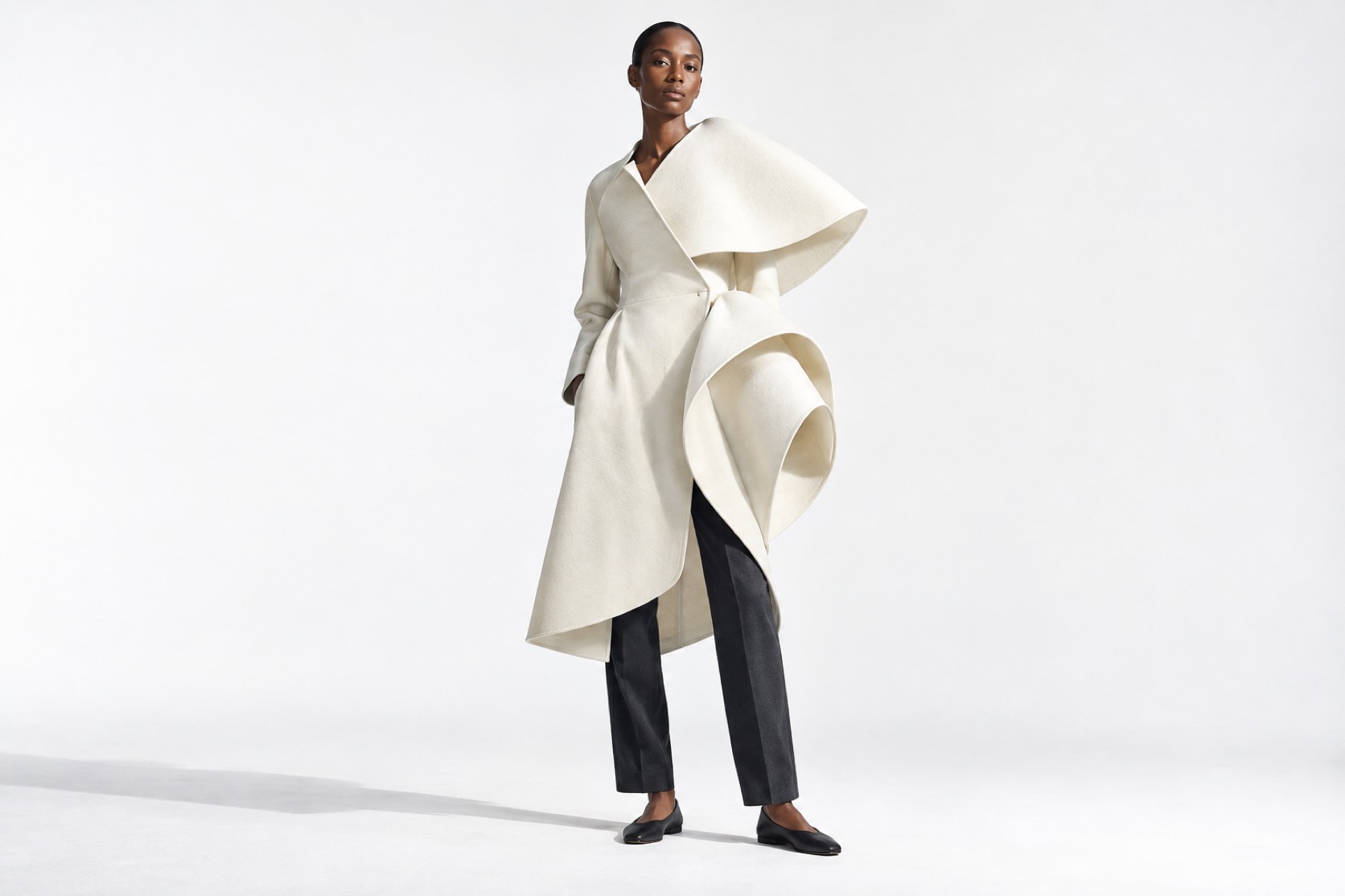 A model wears an ivory coat with sculptural folded volumes and charcoal trousers.