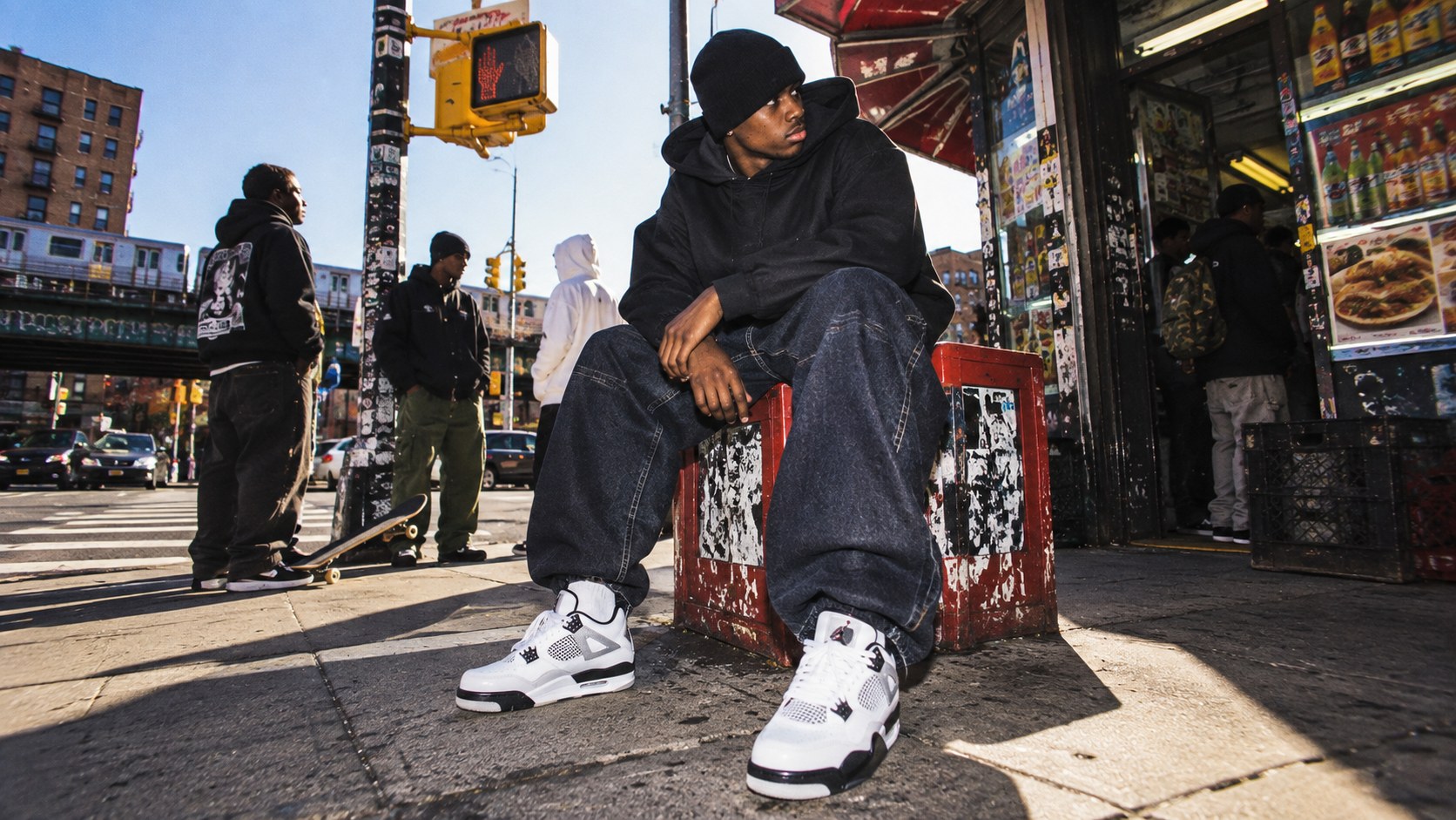 A young man in a black hoodie, baggy dark jeans and white sneakers sits on a red crate outside a New York deli, friends around him on the corner.