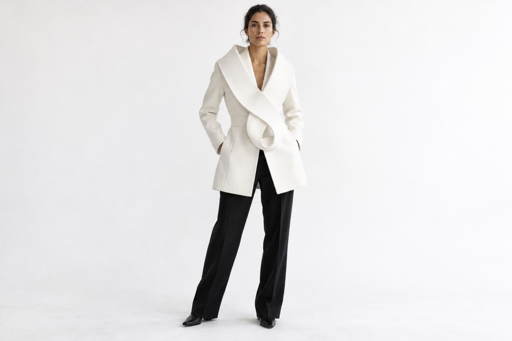 A model wears a white coat with a looping sculptural lapel and black trousers.